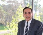 Ein Mann in Anzug und Krawatte sitzt drinnen vor einem großen Fenster. Grüne Bäume und Sonnenlicht bilden eine ruhige Kulisse. Er blickt mit einem subtilen Lächeln in die Kamera und strahlt die selbstbewusste Ausstrahlung eines Menschen aus, der bereit für seine nächste Vorlesung an der Hochschule Coburg ist.