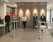 Eine Gruppe von sieben Personen, die mit der Hochschule Coburg verbunden sind, steht mit Abstand in einem Innenraum mit modernem Dekor. Sie tragen formelle Kleidung und sitzen um kleine runde Tische herum, mit Blick in die Kamera. Im Hintergrund sind eine beleuchtete Wand und große Fenster auf der rechten Seite zu sehen.