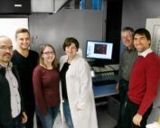 Eine Gruppe von sieben Personen steht in einem Labor der Hochschule Coburg und lächelt in die Kamera. Eine Person trägt einen weißen Laborkittel. Im Hintergrund zeigt ein Computermonitor wissenschaftliche Daten an, während verschiedene Laborgeräte und ein Periodensystem zu sehen sind.