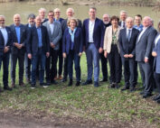 Eine Gruppe von Menschen in formeller Kleidung steht im Freien auf einer Wiese neben einem Fluss und posiert neben einem blauen Schild mit der Aufschrift „Hightech Agenda Bayern“. Mit Bäumen und dem Flussufer im Hintergrund verleihen Vertreter der Hochschule Coburg der Szene ihre Präsenz.