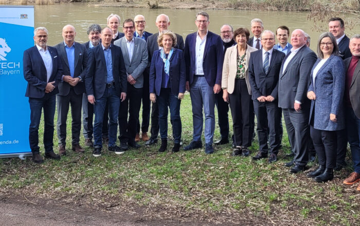 Eine Gruppe von Menschen in formeller Kleidung steht im Freien auf einer Wiese neben einem Fluss und posiert neben einem blauen Schild mit der Aufschrift „Hightech Agenda Bayern“. Mit Bäumen und dem Flussufer im Hintergrund verleihen Vertreter der Hochschule Coburg der Szene ihre Präsenz.