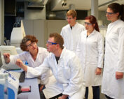Eine Gruppe von fünf Wissenschaftlern der Hochschule Coburg, in Laborkittel und Schutzbrillen gekleidet, versammelt sich im Labor um einen Computerbildschirm. Eine Person zeigt auf den Bildschirm, während die anderen aufmerksam zuschauen, was eine gemeinsame Diskussion oder Analyse nahelegt.