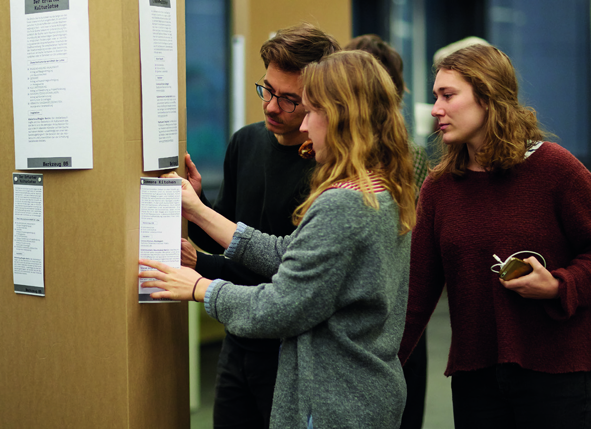 Drei Personen stehen vor einer Schautafel an der Hochschule Coburg, lesen und diskutieren den Text darauf. Sie wirken engagiert und konzentriert auf die präsentierten Informationen. Die Umgebung erinnert an eine Ausstellung oder einen Bildungsraum.