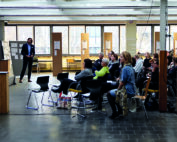 Eine Gruppe von Menschen sitzt im Innenbereich der Hochschule Coburg und hört aufmerksam einer Präsentation zu. Eine Person steht vorne, während eine andere neben einer Tafel mit Unterlagen steht. Der Raum verfügt über große Fenster und einen Fliesenboden, was für eine spannende akademische Atmosphäre sorgt.