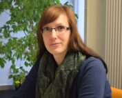 Eine Person mit langen braunen Haaren und Brille sitzt in einem Innenbereich neben einer Blattpflanze. Sie trägt einen dunklen Schal und ein blaues Oberteil. Im Hintergrund sind ein Fenster, ein Teil eines Heizkörpers und ein Emblem der Hochschule Coburg zu sehen.