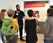 Eine Gruppe von Menschen steht im Halbkreis um einen Mann, der an der Hochschule Coburg ein Poster mit dem Titel „Forschendes Lernen in digitalen Teams“ präsentiert. Sie diskutieren in einem Innenraum. Der Mann gestikuliert, während er erklärt.