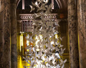 Eine Ausstellung zahlreicher weißer Papiervögel ist in einer hohen, eleganten Spirale in der Hochschule Coburg angeordnet. Diese befindet sich in einem prachtvollen Gebäude mit Marmorsäulen und sanft beleuchteten Bogenfenstern im Hintergrund.