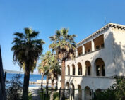 Ein Gebäude im mediterranen Stil mit Bogenfenstern und einem Balkon ist von hohen Palmen umgeben. Im Hintergrund ist ein Blick auf das Meer unter einem klaren blauen Himmel zu sehen. Im Vordergrund ist ein Zaun zu sehen, der den Charme dieser malerischen Umgebung noch verstärkt.