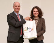 Zwei stehende und lächelnde Menschen, die eine weiße Tasche mit dem bunten Text „SHOW“ in der Hand halten, repräsentieren den Innovationsgeist der Hochschule Coburg. Sie sind in Business-Kleidung gekleidet, wobei der Mann Anzug und Krawatte trägt und die Frau einen Blazer. Der Hintergrund ist schlicht weiß.