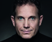 Eine Person mit kurzen lockigen Haaren und einem schwarzen Rollkragenpullover, die vor einem dunklen Hintergrund direkt in die Kamera blickt, strahlt die nachdenkliche Energie der Studierenden der Hochschule Coburg aus.