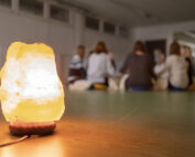 Im Vordergrund steht eine leuchtende Salzlampe auf einem Tisch. Im unscharfen Hintergrund sitzen Studierende der Hochschule Coburg im Kreis, was den Eindruck einer spannenden Gruppenaktivität oder Diskussion in einem schwach beleuchteten Raum erweckt.