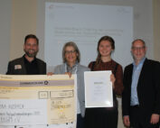 Vier Menschen stehen lächelnd nebeneinander an der Hochschule Coburg. Die Person links hält einen großen Scheck über 1.000 Euro in den Händen, eine andere zeigt stolz eine Urkunde. Im Hintergrund ist eine Präsentationsfolie zu sehen, die auf Neurofeedback-Training verweist.