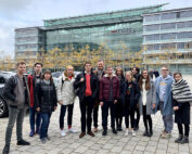 Eine Gruppe von Menschen, möglicherweise Studenten der Hochschule Coburg, steht vor dem Audi-Gebäude und lächelt in die Kamera. Die Kulisse ist eine moderne Plaza mit Bäumen und einer mehrstöckigen Glas- und Stahlkonstruktion im Hintergrund. Alle sind in Winterkleidung gekleidet.