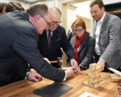 An der Hochschule Coburg versammelt sich eine Gruppe von Menschen in formeller Kleidung um einen Tisch und bastelt kleine Holzkonstruktionen. Ein Mann benutzt einen Hammer, um die Teile zusammenzusetzen, während andere zuschauen und assistieren. Der Tisch ist mit Modellteilen und Werkzeugen übersät, was Teamarbeit und Kreativität in einem akademischen Umfeld fördert.
