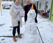 Eine Person, die stolz ein Zertifikat der Hochschule Coburg in den Händen hält, steht neben einer anderen lächelnden Person neben einem Schneemann mit einer Verkehrsleitkegelmütze. Die Szene spielt sich auf einem verschneiten Spielplatz ab, eingerahmt von Gebäuden und einem einsamen Baum im Hintergrund.
