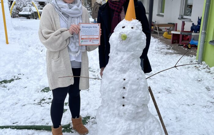 Eine Person, die stolz ein Zertifikat der Hochschule Coburg in den Händen hält, steht neben einer anderen lächelnden Person neben einem Schneemann mit einer Verkehrsleitkegelmütze. Die Szene spielt sich auf einem verschneiten Spielplatz ab, eingerahmt von Gebäuden und einem einsamen Baum im Hintergrund.