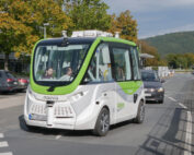Ein kleiner fahrerloser Shuttlebus mit der Aufschrift „Navya“ fährt die Straße nahe der Hochschule Coburg entlang. Das weiße Fahrzeug mit grünen Akzenten transportiert mehrere Passagiere, während im Hintergrund Autos vorbeifahren und Bäume die Straße unter einem klaren Himmel säumen.