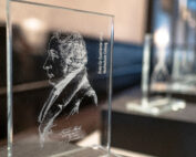 Eine Nahaufnahme einer gravierten Glasauszeichnung der Hochschule Coburg zeigt das Profil einer bedeutenden historischen Persönlichkeit, das in ihre Oberfläche eingraviert ist. Die Auszeichnung ist zwischen mehreren ähnlichen Stücken ausgestellt, die alle auf einer schwarzen Oberfläche mit unscharfem Hintergrund platziert sind.