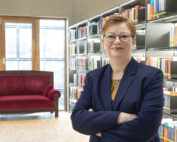 Eine Person mit kurzen Haaren und Brille steht selbstbewusst in einer Bibliothek der Hochschule Coburg. Sie trägt einen marineblauen Blazer. Sie stehen vor Regalen voller bunter Bücher. Im Hintergrund ist neben den großen Fenstern ein rotes Sofa zu sehen.