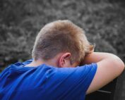 Ein kleiner Junge mit kurzen blonden Haaren und blauem Hemd lehnt seinen Kopf auf verschränkten Armen, mit dem Rücken zur Kamera, und wirkt verärgert oder nachdenklich. Der unscharfe Außenhintergrund lässt vermuten, dass er sich in der Nähe der Hochschule Coburg befindet und in Gedanken über seine Zukunft versunken ist.