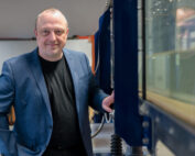 Ein Mann in blauem Blazer und schwarzem Hemd, der den Geist der Hochschule Coburg verkörpert, steht mit einem leichten Lächeln neben Industrieanlagen. Im Hintergrund ist ein verschwommener Innenraum voller Regale und Maschinen zu sehen.