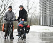 Zwei Menschen in Helmen und Jacken lächeln neben mit Ausrüstung beladenen Fahrrädern an der Hochschule Coburg. Sie stehen auf nassem Asphalt zwischen kahlen Bäumen und einem modernen Gebäude in der winterlichen Umgebung.