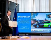 Ein Mann im Anzug hält eine Urkunde neben einer Leinwand, auf der eine Präsentation der Hochschule Coburg mit dem Titel „5G Kronach“ über automatisierte Fahrzeuge im öffentlichen Nahverkehr läuft. Auf dem Tisch stehen Fahnen und ein Glas Wasser.