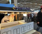 Mitten im Trubel der Veranstaltung inspiziert eine Person mit langen blonden Haaren ein detailreiches Architekturmodell eines modernen Gebäudes. Darüber leuchtet das Schild „Designfabrik“ und weist auf die Verbindung zur Hochschule Coburg hin. Besucher und Kleiderständer sorgen für eine lebendige Kulisse.