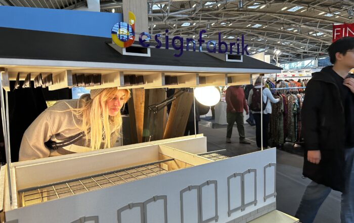 Mitten im Trubel der Veranstaltung inspiziert eine Person mit langen blonden Haaren ein detailreiches Architekturmodell eines modernen Gebäudes. Darüber leuchtet das Schild „Designfabrik“ und weist auf die Verbindung zur Hochschule Coburg hin. Besucher und Kleiderständer sorgen für eine lebendige Kulisse.