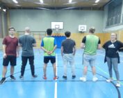 Fünf Personen stehen in einer Turnhalle der Hochschule Coburg vor einem Basketballkorb. Vier tragen Sportkleidung und kehren der Kamera den Rücken zu, während einer rechts nach vorne blickt und selbstbewusst lächelt und die Arme verschränkt. Die Turnhalle ist mit blauem Boden und Holzpaneelen ausgestattet und spiegelt den lebendigen Geist der Coburger Gemeinschaft wider.