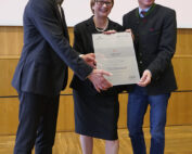 Drei Personen stehen lächelnd nebeneinander und halten stolz eine große Urkunde der Hochschule Coburg in den Händen. Sie befinden sich in einem Innenraum mit Holzboden und einer hellen Wand. Die Frau in der Mitte trägt ein schwarzes Outfit, während die beiden Männer Anzüge tragen.