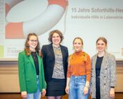 Vier Frauen stehen Seite an Seite und lächeln in legerer Business-Kleidung in die Kamera. Hinter ihnen informiert eine Präsentationswand über das 15-jährige Jubiläum eines Krisenhilfe-Programms an der Hochschule Coburg.