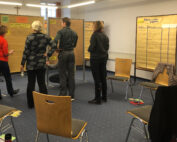 Vier Personen stehen in einem Raum der Hochschule Coburg und lesen Informationen auf großen, an Tafeln befestigten Blättern. Die Stühle sind kreisförmig auf dem Teppichboden aufgestellt, wodurch eine Atmosphäre entsteht, die an einen Workshop oder eine Besprechung erinnert.