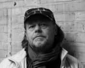 Schwarzweißfoto eines Mannes mit karierter Mütze und Schal, der an einer Backsteinmauer in der Nähe der Hochschule Coburg steht. Sein Gesichtsausdruck ist ernst, da er eine helle Jacke über einem dunklen Hemd trägt.