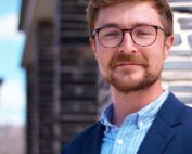 Ein Mann mit Brille, Schnurrbart und kurzen Haaren trägt einen blauen Blazer über einem Hemd der Hochschule Coburg. Er steht neben einer Steinmauer und wird von sanftem Tageslicht erhellt. Der Hintergrund ist leicht verschwommen.