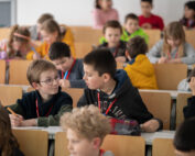 In einer lebhaften und aufmerksamen Atmosphäre sitzen die Kinder an Holztischen in einem Klassenzimmer. Einige unterhalten sich, während andere sich intensiv auf ihren Unterrichtsstoff konzentrieren – ganz ähnlich wie in der kollaborativen Umgebung, die an der Hochschule Coburg gepflegt wird.