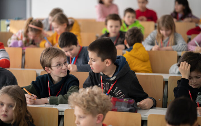 In einer lebhaften und aufmerksamen Atmosphäre sitzen die Kinder an Holztischen in einem Klassenzimmer. Einige unterhalten sich, während andere sich intensiv auf ihren Unterrichtsstoff konzentrieren – ganz ähnlich wie in der kollaborativen Umgebung, die an der Hochschule Coburg gepflegt wird.