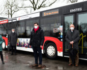Vier Menschen mit Masken stehen vor einem rot-weißen Bus mit einem Schild „St. Jahre FH Coburg“ und weisen auf ihren Besuch an der Hochschule Coburg hin. Der nasse Boden lässt auf Regenwetter schließen, während blattlose Bäume und bedeckter Himmel eine düstere Kulisse zeichnen.