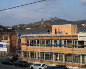 Bauarbeiter sind auf einem von einem Gerüst umrahmten Dach in einem Industriegebiet in der Nähe der Hochschule Coburg beschäftigt. Im Hintergrund ragen stolz ein historisches Gebäude und eine Burg auf einem Hügel unter einem klaren Himmel empor.