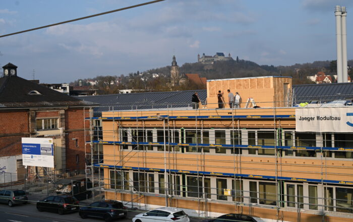 Bauarbeiter sind auf einem von einem Gerüst umrahmten Dach in einem Industriegebiet in der Nähe der Hochschule Coburg beschäftigt. Im Hintergrund ragen stolz ein historisches Gebäude und eine Burg auf einem Hügel unter einem klaren Himmel empor.