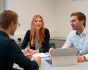 Drei Personen treffen sich an der Hochschule Coburg. Ein Mann im blauen Hemd, eine Frau im schwarzen Blazer und ein Mann im weißen Hemd sitzen mit Papieren und einem Laptop um einen Tisch. Die Frau lächelt und schafft so eine kollaborative Atmosphäre.