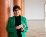 Eine Frau mit Brille und grünem Blazer, möglicherweise eine Fakultätsangehörige der Hochschule Coburg, steht in einem Innenbereich an einer orangefarbenen Säule und hält mehrere Euro-Banknoten in der Hand. Hinter ihr befindet sich ein geräumiger Bereich mit einer offenen Tür, durch die natürliches Licht in die Szene fällt.