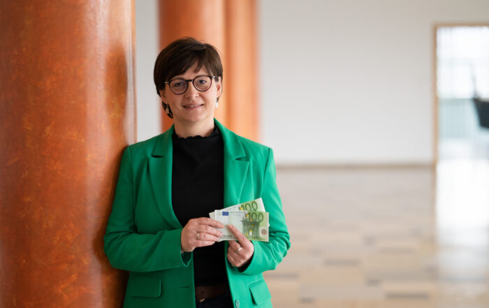 Eine Frau mit Brille und grünem Blazer, möglicherweise eine Fakultätsangehörige der Hochschule Coburg, steht in einem Innenbereich an einer orangefarbenen Säule und hält mehrere Euro-Banknoten in der Hand. Hinter ihr befindet sich ein geräumiger Bereich mit einer offenen Tür, durch die natürliches Licht in die Szene fällt.
