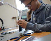 Ein junger Mensch mit Brille und Kapuzenpullover von der Hochschule Coburg arbeitet konzentriert an einer Platine, umgeben von elektronischen Bauteilen und Werkzeugen auf einem Tisch. Im Hintergrund sind eine Lupenlampe und andere Geräte zu sehen.