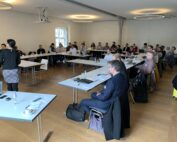 Eine Gruppe von Menschen sitzt in einem Konferenzraum der Hochschule Coburg, ganz vorne steht eine Frau, die vermutlich einen Vortrag hält. Die Tische sind U-förmig angeordnet, die Anwesenden hören aufmerksam zu.