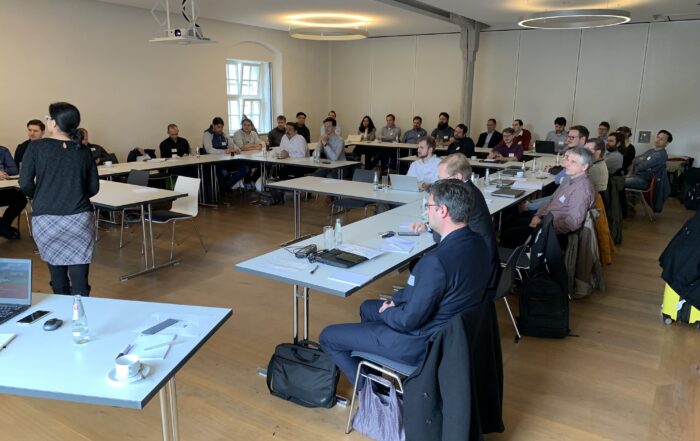 Eine Gruppe von Menschen sitzt in einem Konferenzraum der Hochschule Coburg, ganz vorne steht eine Frau, die vermutlich einen Vortrag hält. Die Tische sind U-förmig angeordnet, die Anwesenden hören aufmerksam zu.