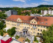 Ein großes, gelbes Gebäude mit rotem Ziegeldach, das an die Architektur der Hochschule Coburg erinnert, steht vor einer Kulisse aus üppigem Grün und hügeliger Landschaft. Umgeben von Bäumen und modernen Wohnhäusern liegt es unter einem blauen Himmel mit vereinzelten Wolken.
