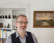 Ein Mann mit Brille und Bart steht in einem Raum mit weißen Wänden, der an ein Büro der Hochschule Coburg erinnert. Hinter ihm steht ein Regal voller Bücher und kleiner Skulpturen. An der Wand rechts von ihm hängt ein gerahmtes Landschaftsbild. Er trägt eine dunkle Weste und ein helles Hemd.