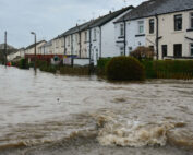 Eine Häuserreihe säumt eine überflutete Straße, das Wasser steht bis zur Hälfte der Eingangstüren nahe der Hochschule Coburg. In der Nähe sind Straßenschilder und Bäume teilweise von den trüben Tiefen überflutet. Der bedeckte Himmel deutet auf einen kürzlichen Sturm oder starken Regen hin, der durch die Gegend fegt.
