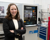 Eine Frau mit langen braunen Haaren, einem schwarzen Blazer und einer weißen Bluse steht lächelnd vor einem Concept Laser 3D-Drucker an der Hochschule Coburg. Das Labor ist mit verschiedenen technischen Geräten und Schränken ausgestattet, die den Spitzentechnologiestandard der Hochschule widerspiegeln.